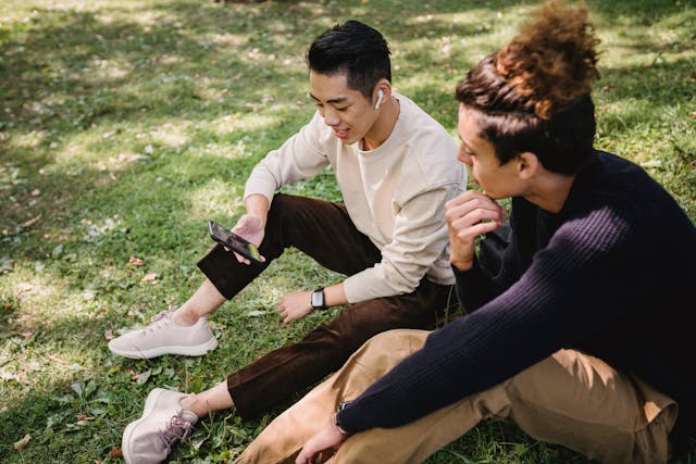 Zwei männliche Freunde sehen sich im Gras sitzend gemeinsam ein Video auf einem Smartphone an.