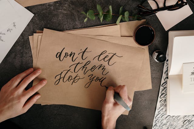 Someone writing, “Don’t let them stop you,” in a beautiful cursive font on brown paper with a brush pen.