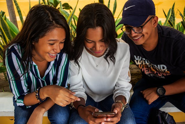 Três adolescentes a sorrir enquanto olham juntos para uma publicação nas redes sociais através do telemóvel.