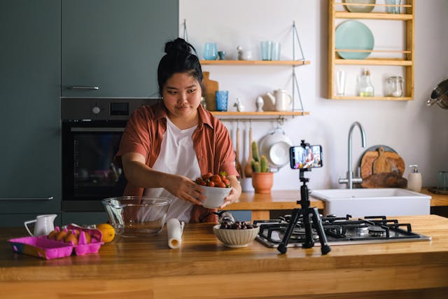 Uma influenciadora gastronómica a gravar um vídeo de uma receita passo a passo na cozinha com o telemóvel e um tripé.