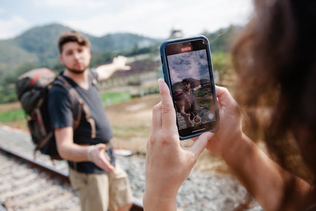 Uma mulher a filmar um vídeo de um viajante do sexo masculino ao ar livre com o seu telemóvel.
