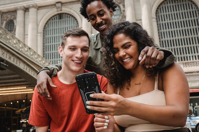 Três amigos a sorrir e a rir enquanto vêem algo no telemóvel.