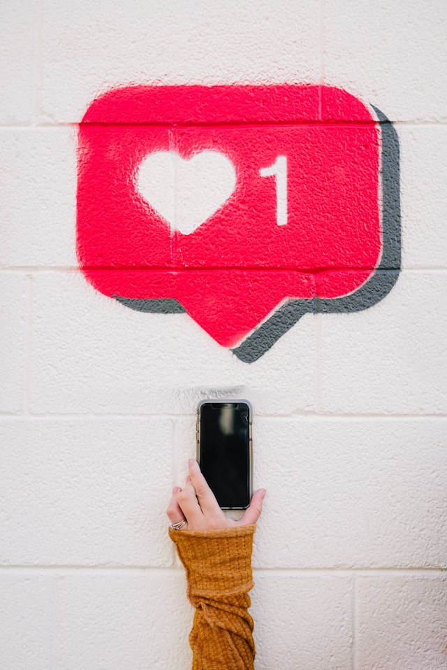 A mão de alguém a segurar um smartphone com o símbolo "like" de Instagram pintado por cima, numa parede.