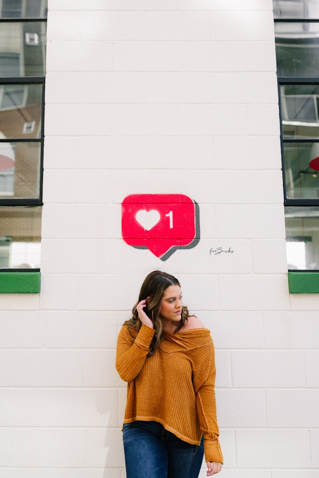O femeie își ascunde părul după ureche în timp ce pozează în fața unui perete pe care este pictat simbolul Instagram Like.