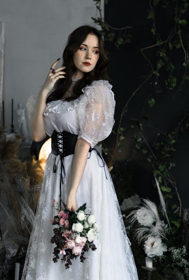 A woman wearing a lacy white dress and a black corset while holding a bouquet of pink, white, and black flowers.
