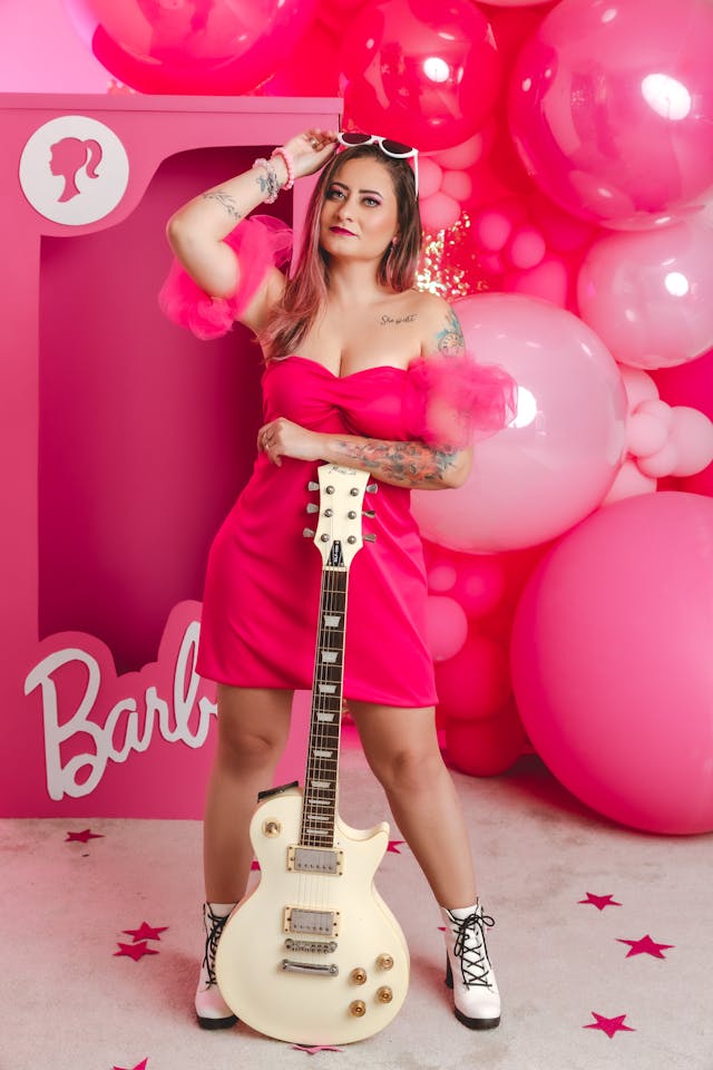 Eine Frau in einem pinkfarbenen Kleid, die eine Gitarre hält und vor pinkfarbenen Luftballons und einer lebensgroßen Barbie-Box steht.