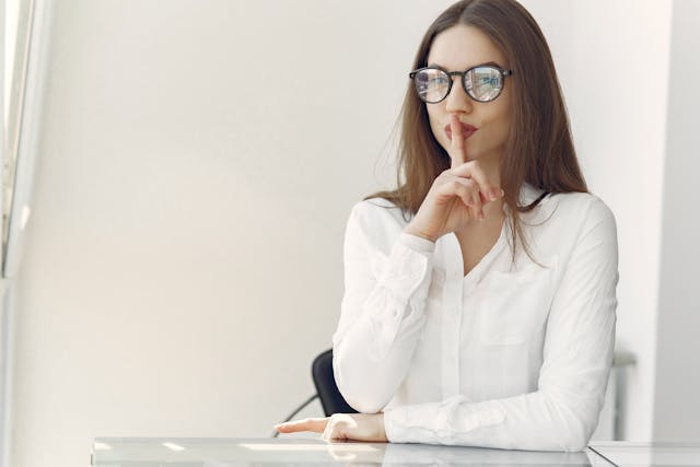 Una donna con occhiali e abbigliamento da ufficio che si preme l'indice sulle labbra con un movimento "shh".