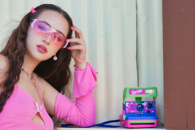 Eine junge Frau mit rosa Kleidung und Sonnenbrille neben einer alten Barbie-Spielzeugkamera.