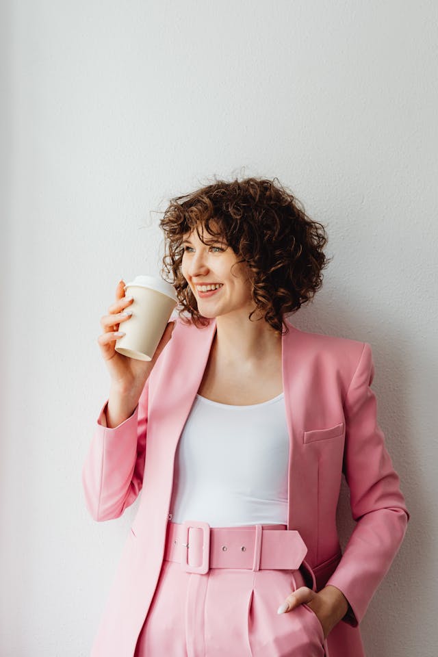 Eine Frau mit lockigem Haar, die einen rosa Anzug trägt, lächelt und eine Tasse Kaffee in der Hand hält.