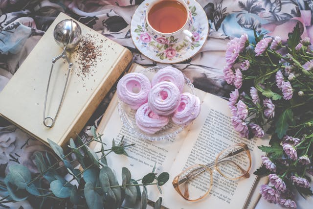 A flat-lay photo of an open book, flowers, pink desserts, reading glasses, and a cup of tea.