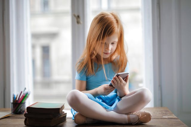 O fetiță cu părul roșcat ține un telefon în timp ce stă pe o masă cu picioarele încrucișate.