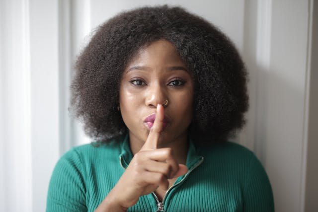 Eine Frau in einem grünen Hemd hält den Finger an die Lippen und macht eine "Pst"-Geste.