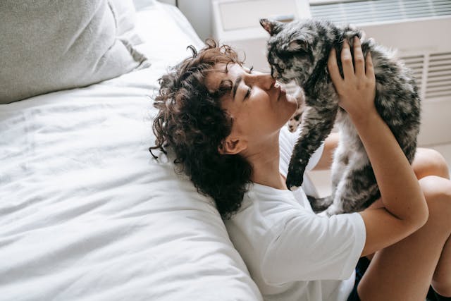 Una donna sdraiata contro un letto mentre tiene in braccio il suo gatto nero e grigio e lo bacia.