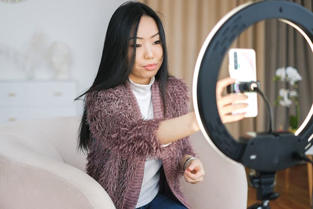 A female influencer in a fuzzy cardigan positioning her phone, tripod, and ring light to record a video of herself.