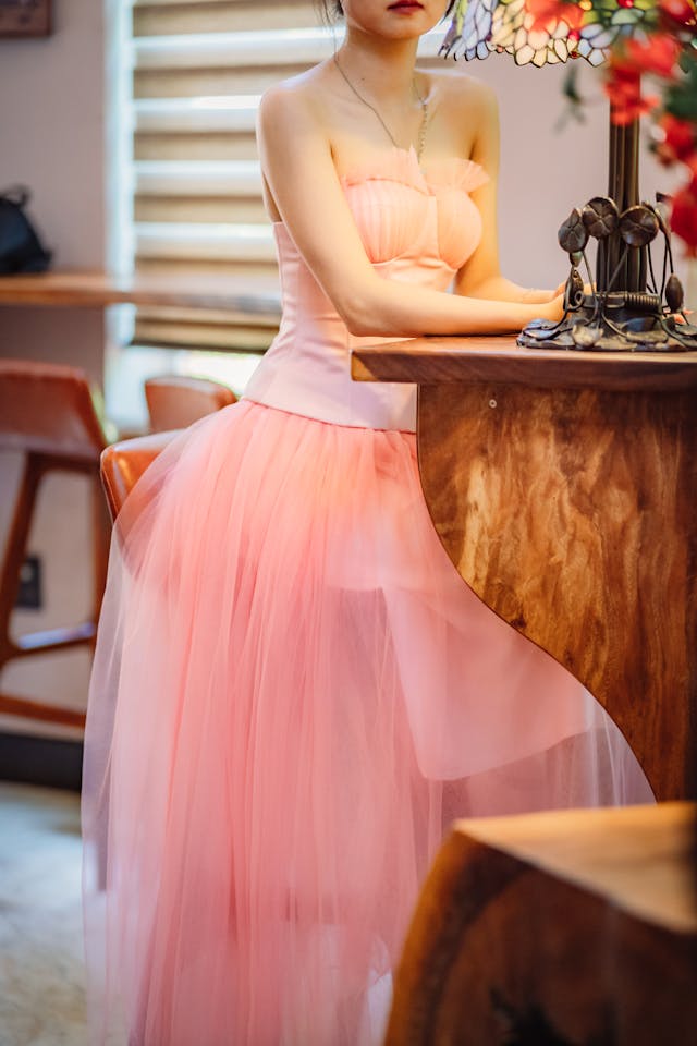 분홍색 얇은 명주 그물 코르셋 드레스에 긴 치마를 입은 여성이 의자에 앉아 있습니다.