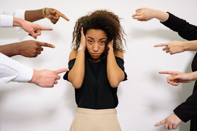 A stressed-looking woman covering her ears while other people point their fingers at her.