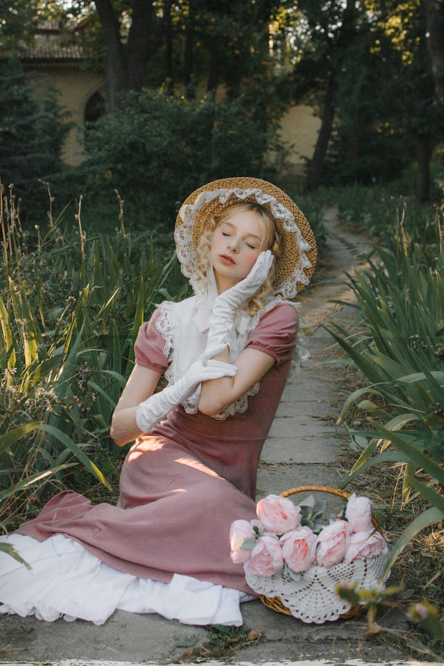빅토리아 시대 드레스와 장갑, 모자를 쓰고 정원에서 눈을 감고 앉아 포즈를 취하고 있는 여성.