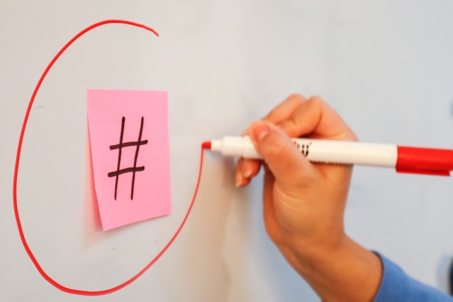 Cineva care folosește un marker roșu pentru a înconjura un bilețel autocolant cu simbolul hashtag lipit pe o tablă albă.