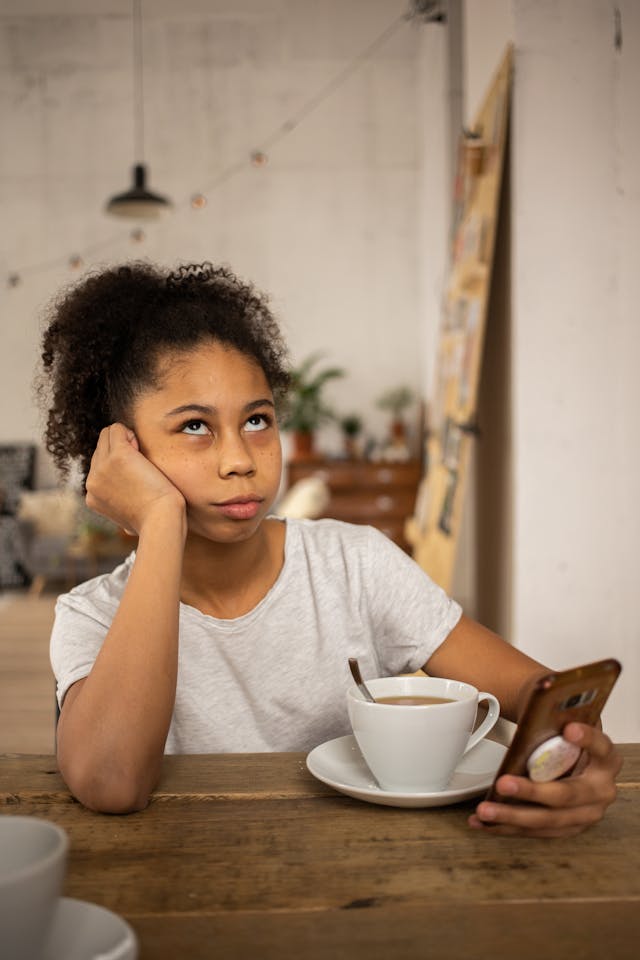 O adolescentă supărată care stă la o masă cu o cafea și își dă ochii peste cap în timp ce dă scroll pe telefon.