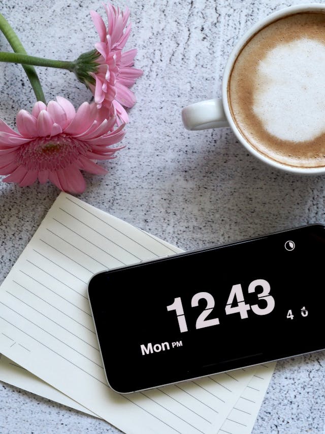  Un iPhone afișează ora curentă pe ecran, lângă niște hârtie, o floare și o ceașcă de cafea pe o masă.